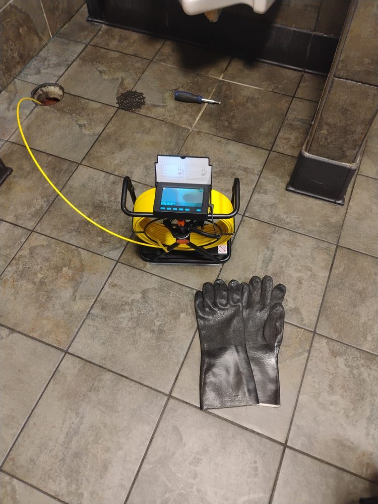 A drain inspection camera with a yellow cable and protective gloves laid out on a tile floor.