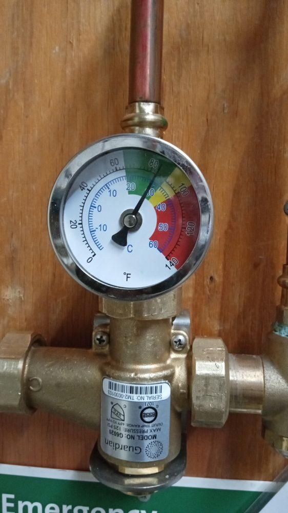 A brass mixing valve on copper piping with a round temperature gauge reading in the safe range.