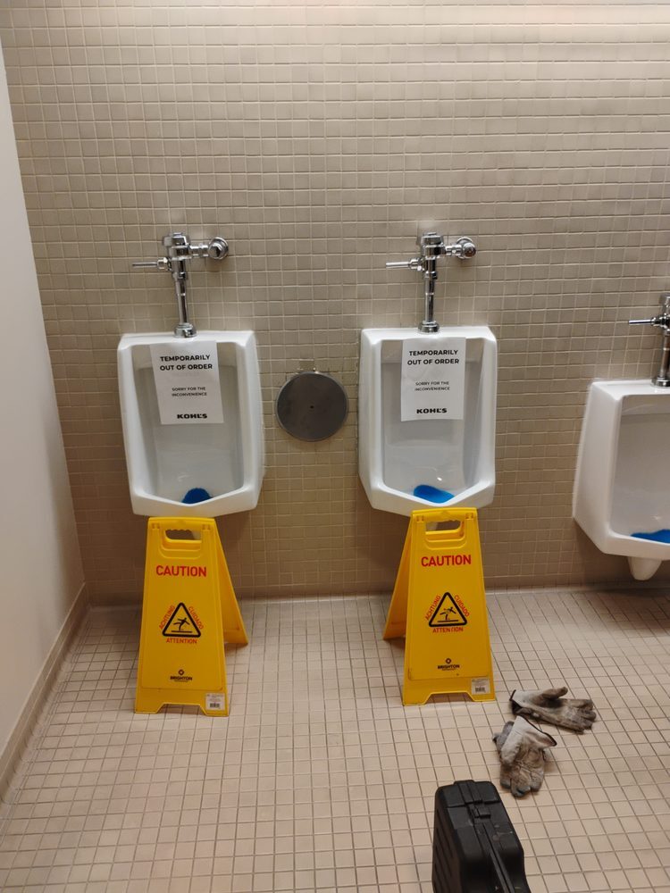 A tiled restroom with two urinals, caution wet floor signs and work gloves set out during maintenance.